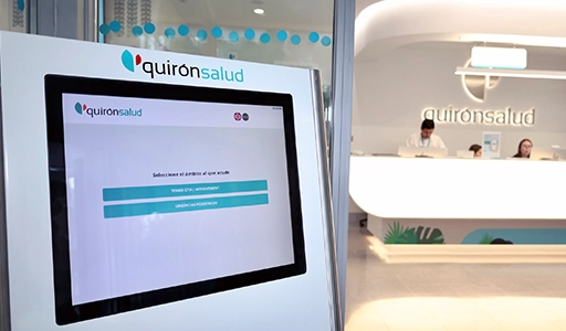 A quirónsalud touchscreen device is visible, with three employees seated at a table behind it (Photo)