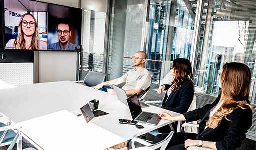 Two women and one man are together in a meeting room; two additional employees are visible on the screen (Photo)